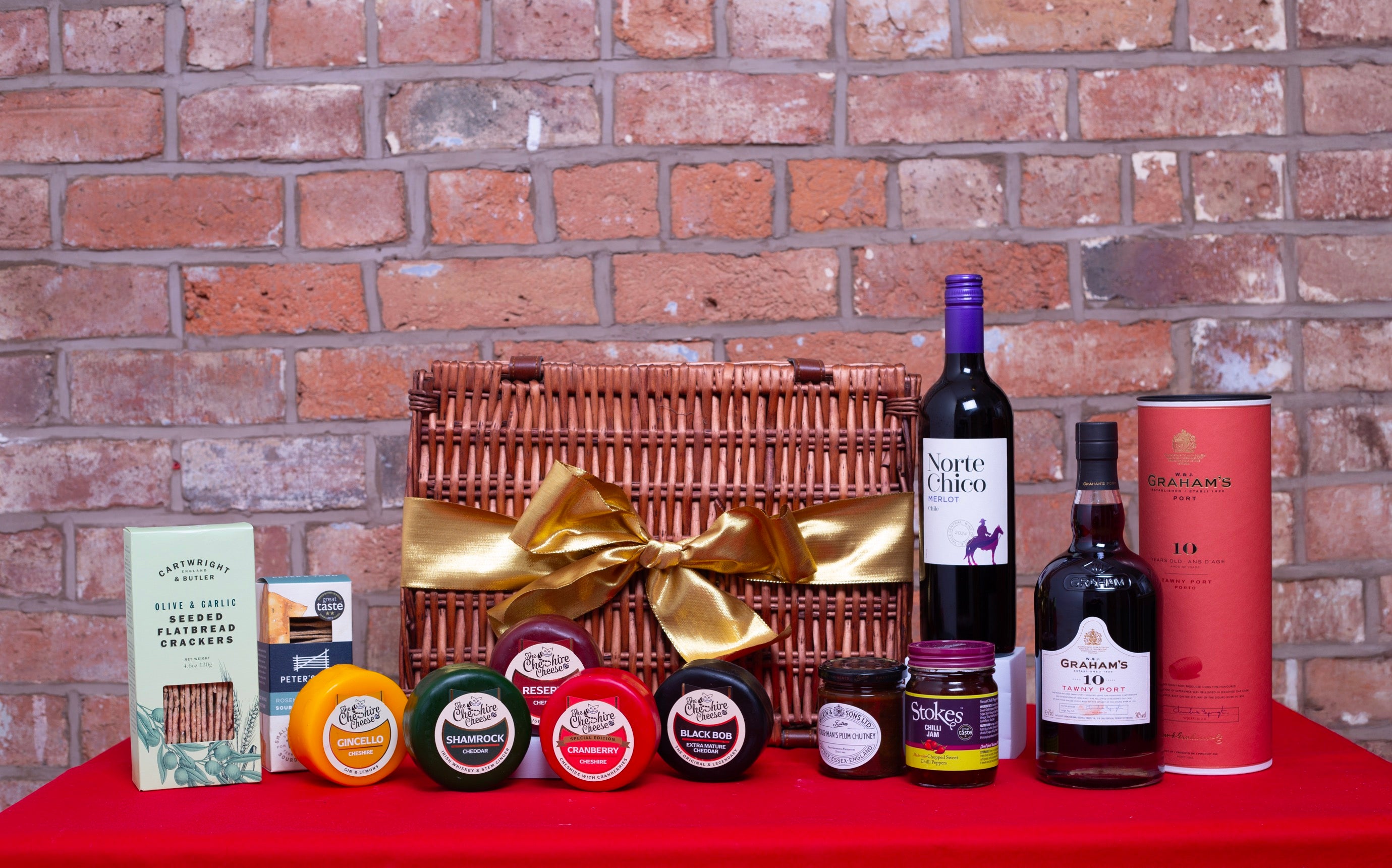 Gourmet gift basket with wine, cheese, and other items against a brick wall.