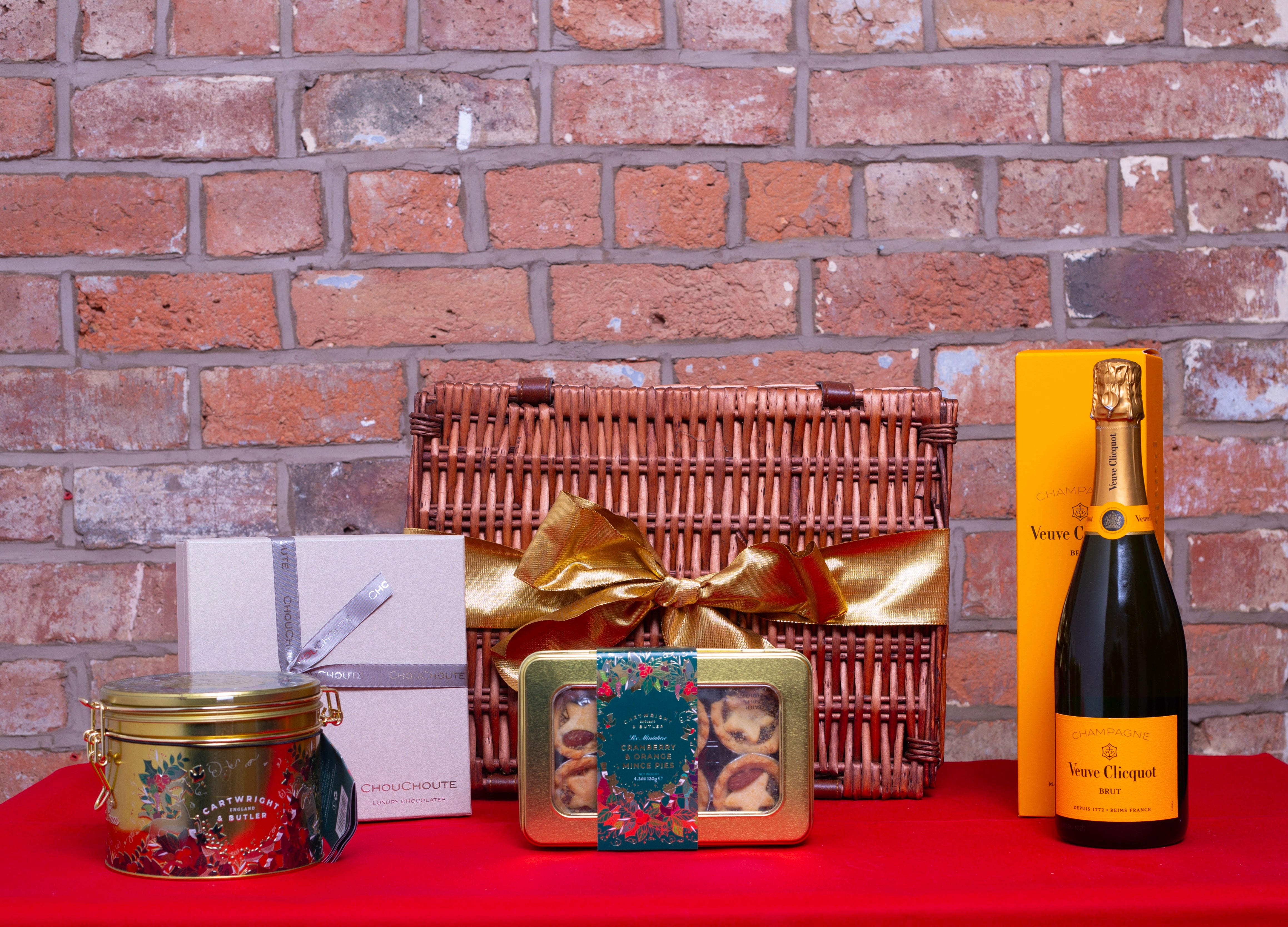 Gift basket with a bottle of champagne, box of chocolates, mince pies, Christmas pudding against a brick wall.
