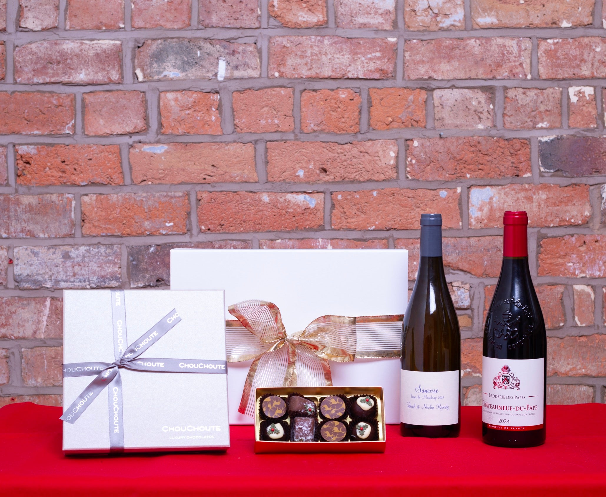 Two bottles of wine, a box of chocolates, and wrapped gifts on a red surface with a brick wall background.